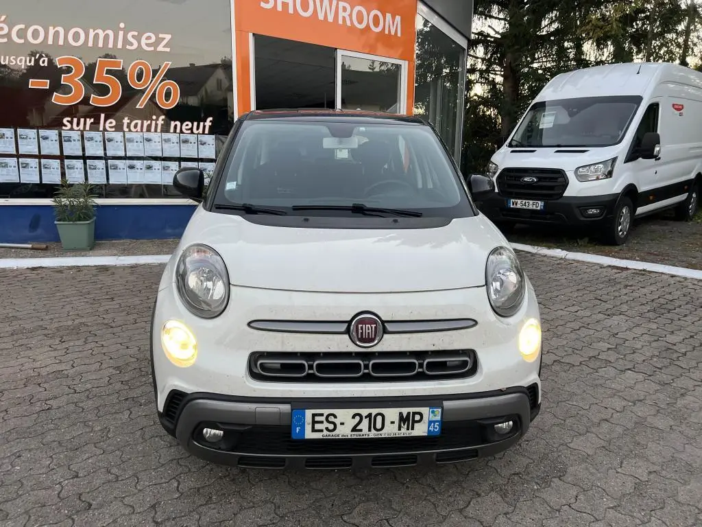 Vue frontale d'une Fiat 500L 1.4i 16V Lounge blanche avec toit noir, feux allumés et logo Fiat visible.