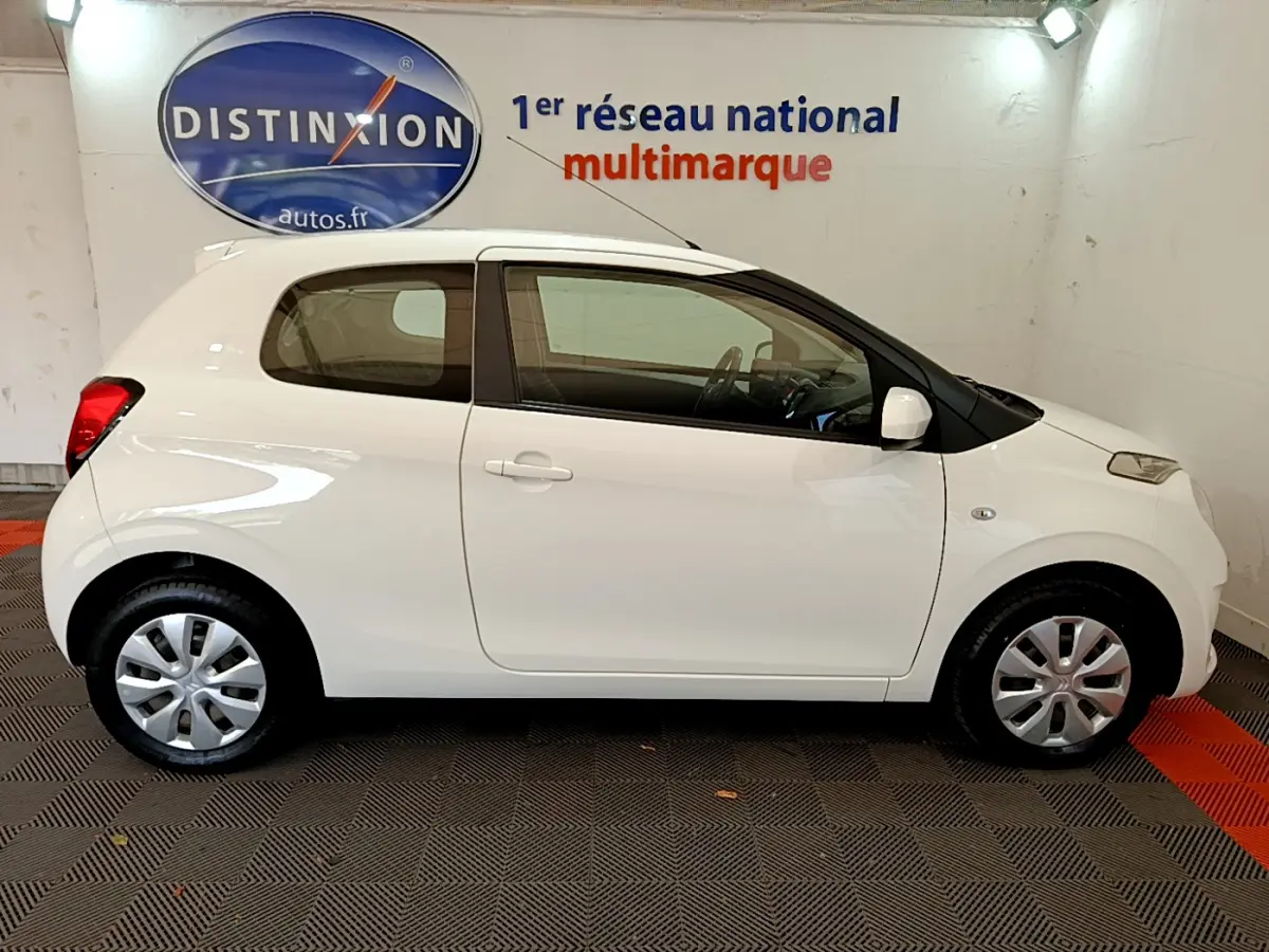 Profil côté droit d'une Citroën C1 blanche 3 portes, avec jantes acier et enjoliveurs, en intérieur showroom.