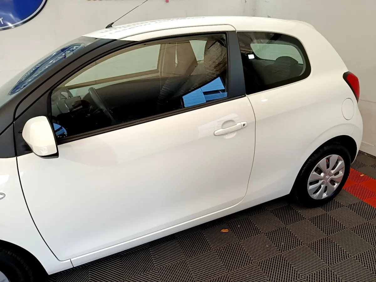 Profil droit d'une Citroën C1 blanche 3 portes, avec rétroviseur et jante acier visibles, sur sol intérieur.