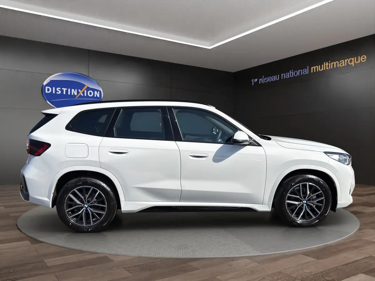 BMW X1 sDrive20d blanc vue 3/4 arrière droit, avec jantes noires et aileron de toit visible.