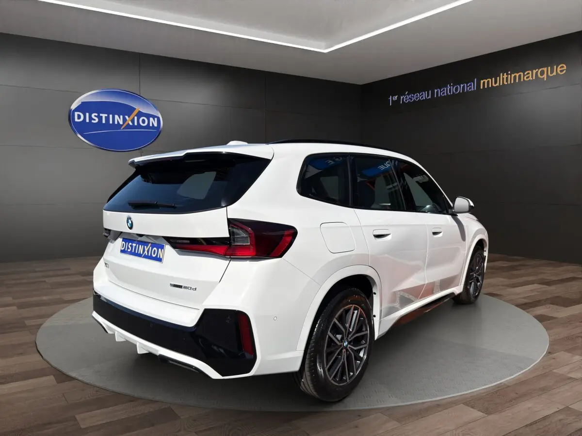 BMW X1 sDrive20d blanc vue 3/4 avant droite avec calandre noire et jantes foncées en showroom