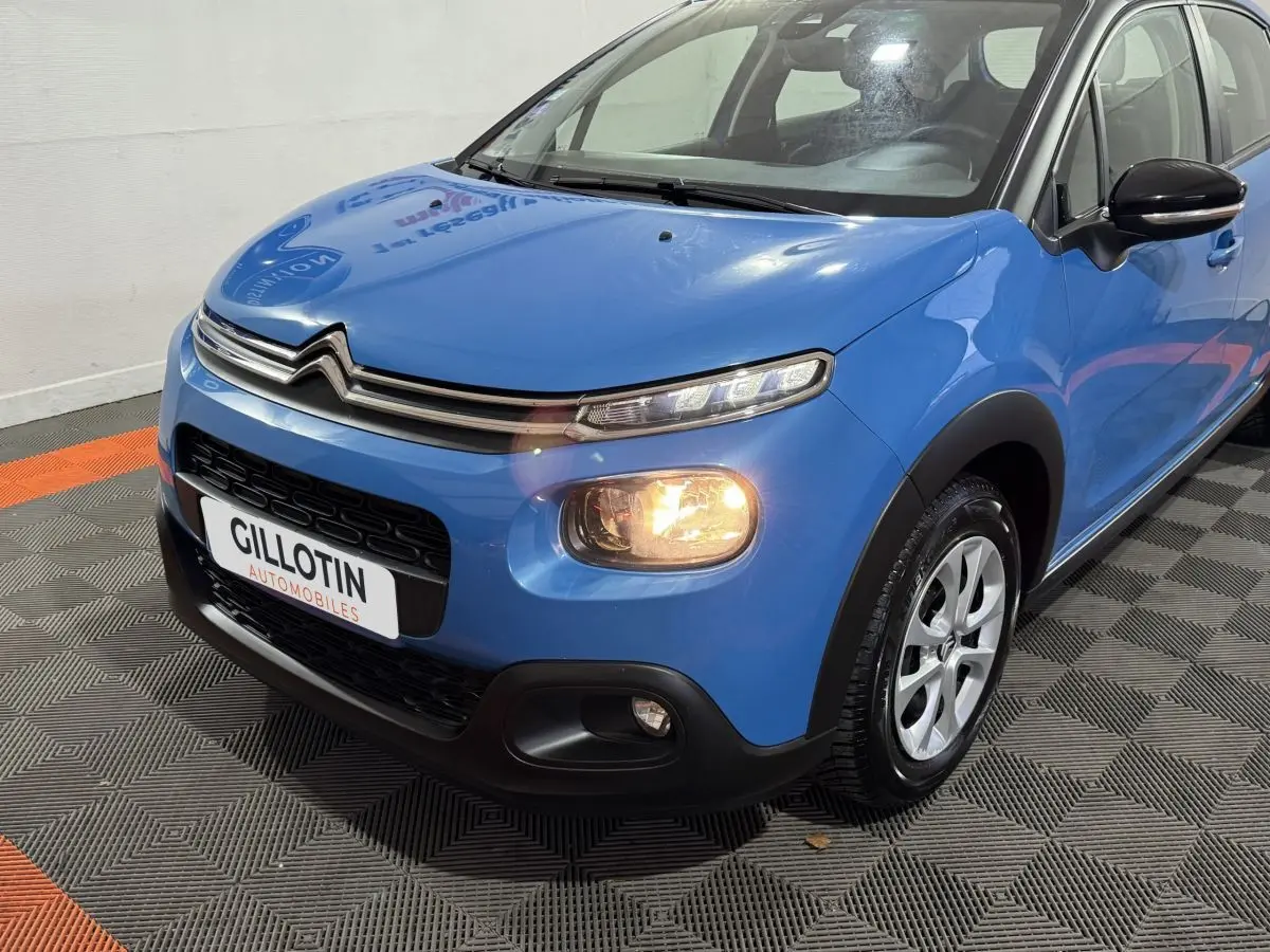 Vue 3/4 avant droit d'une Citroën C3 bleu avec feux allumés et jantes alliage visibles sur sol intérieur.