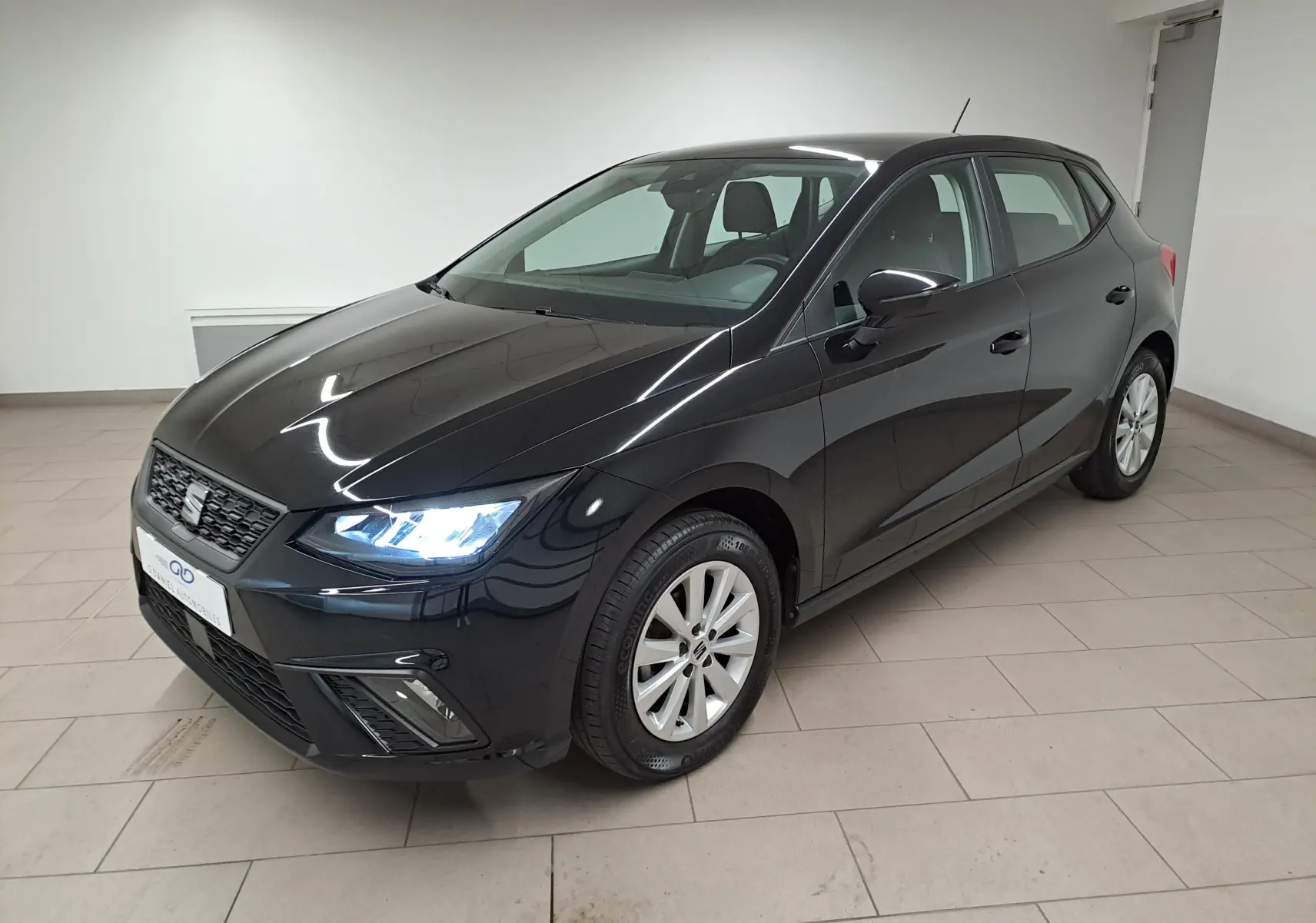 SEAT Ibiza 2024 noir minuit vue 3/4 avant droit, phares allumés et jantes alliage visibles en intérieur showroom.