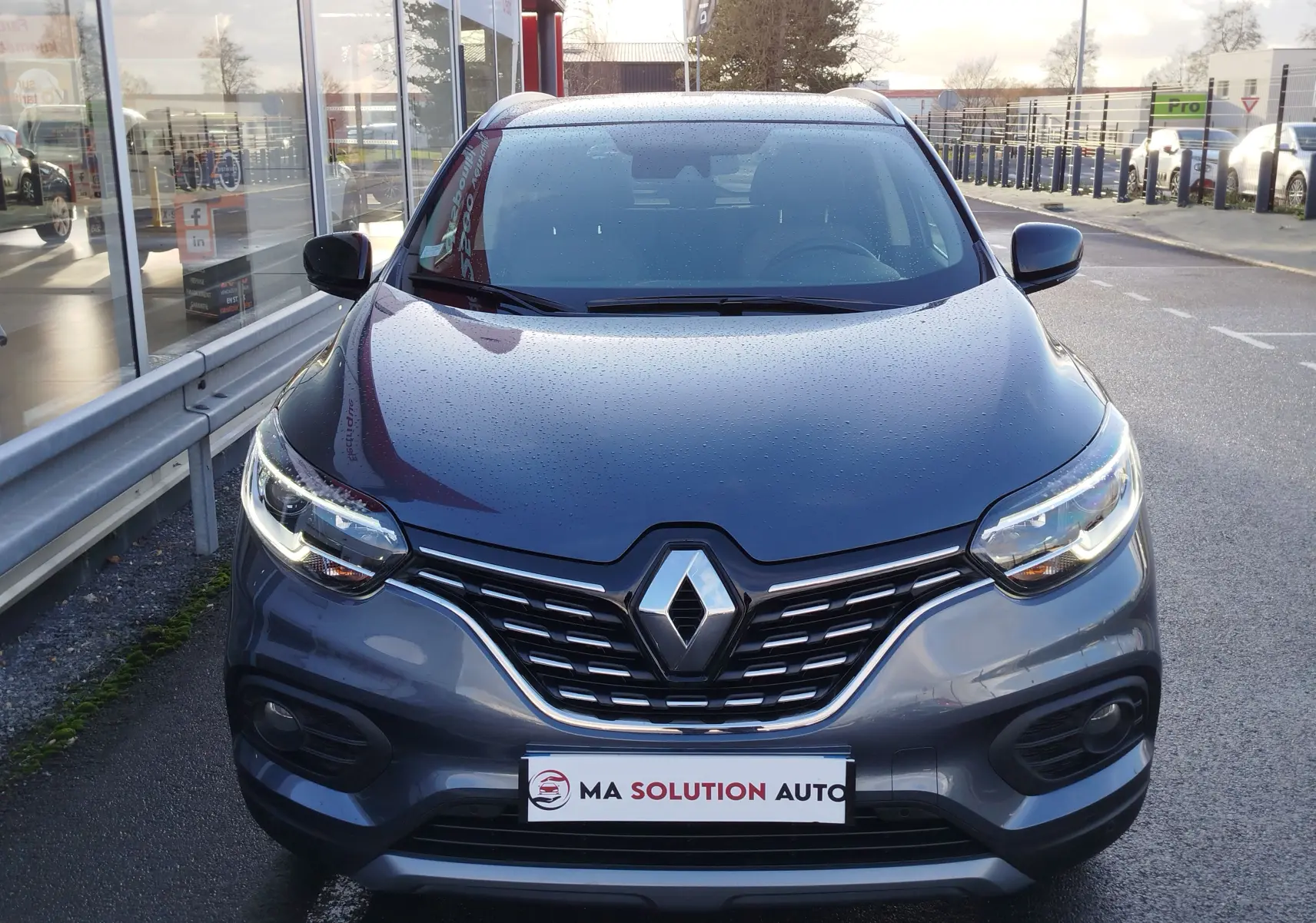 Vue frontale d'un Renault Kadjar gris titanium 2021 avec calandre chromée et feux LED allumés sous la pluie.