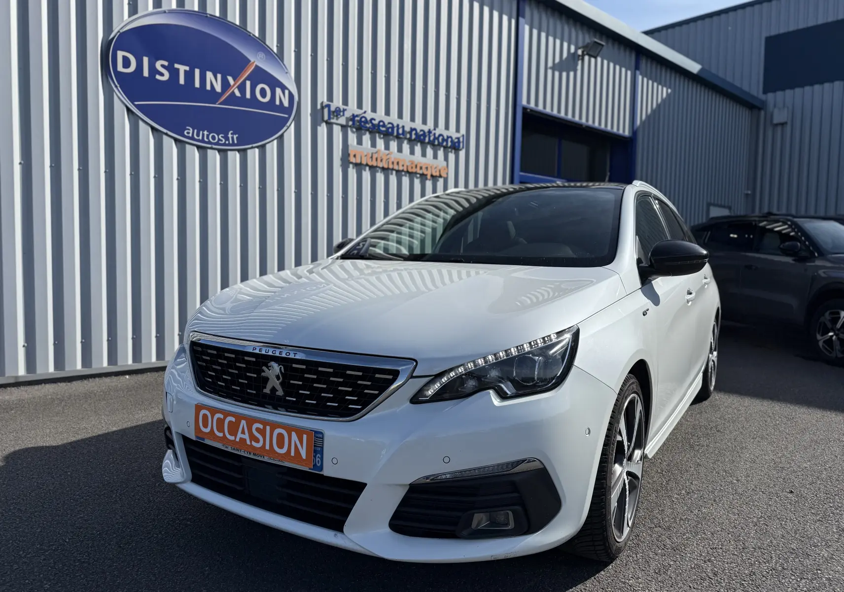 Peugeot 308 SW blanc nacré vue 3/4 avant droit, avec calandre noire et plaque orange "OCCASION" devant un bâtiment industriel.
