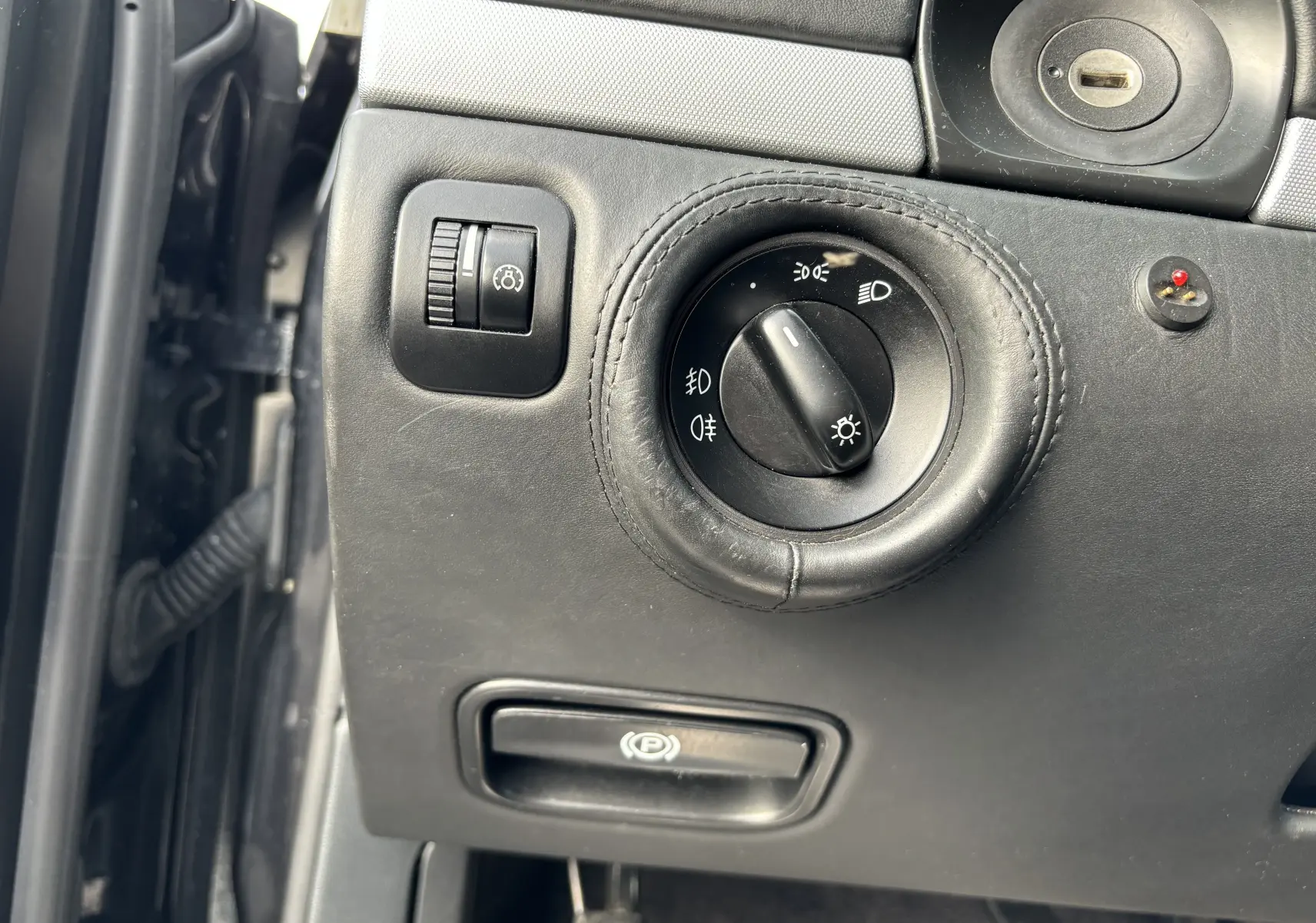 Gros plan sur le tableau de bord noir du Porsche Cayenne 2007, montrant la commande des phares et le frein à main électrique.