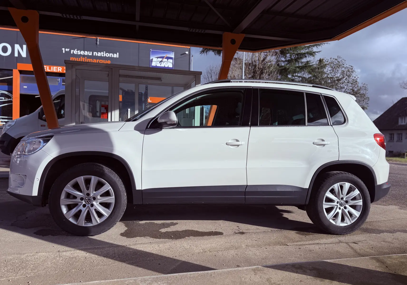 Profil côté gauche d’un Volkswagen Tiguan blanc de 2010, avec jantes alliage et vitres teintées, sous un abri.