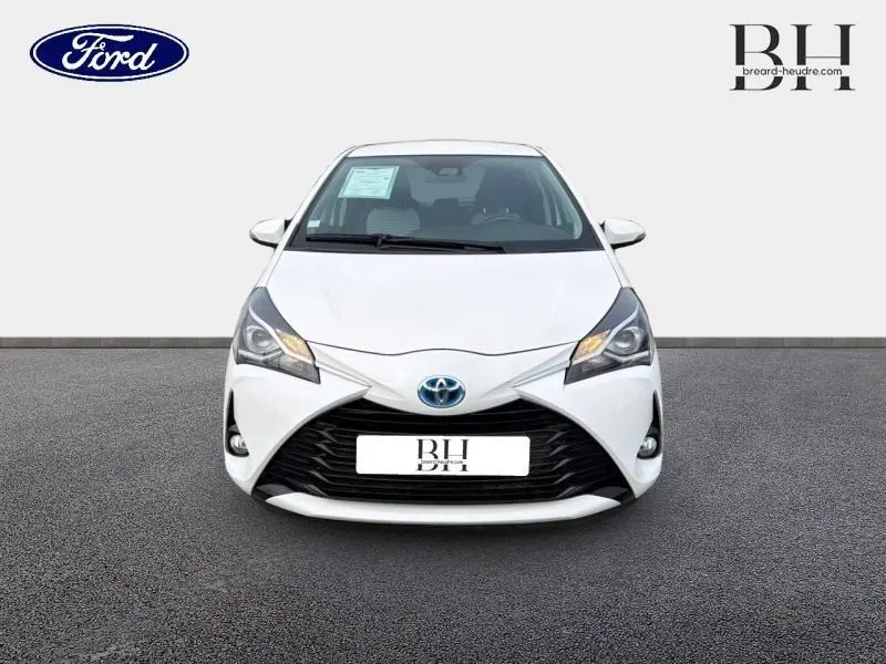 Vue frontale d'une Toyota Yaris 116h France Business 5p blanche avec calandre noire et phares allumés.