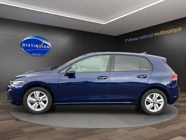 Profil latéral droit d'une Volkswagen Golf VIII bleu foncé, cinq portes, en intérieur showroom avec logo Distinxion en arrière-plan.
