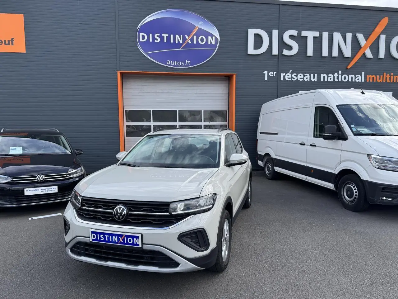 Volkswagen T-Cross gris Ascot en 3/4 avant droit, avec calandre noire et projecteurs LED, devant un garage Distinxion.