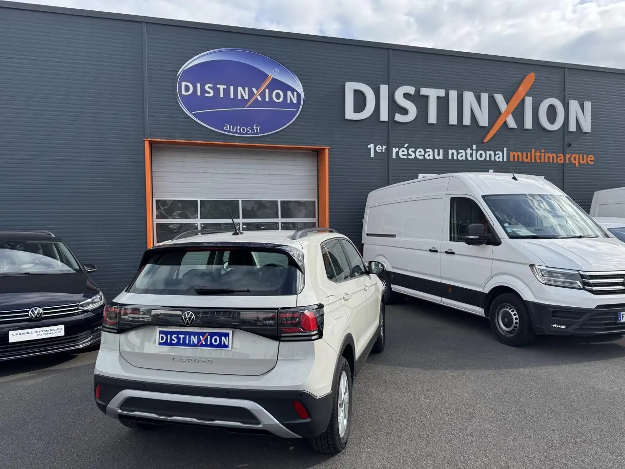 Vue arrière d'un Volkswagen T-Cross gris Ascot garé devant un bâtiment Distinxion avec un utilitaire blanc à côté.