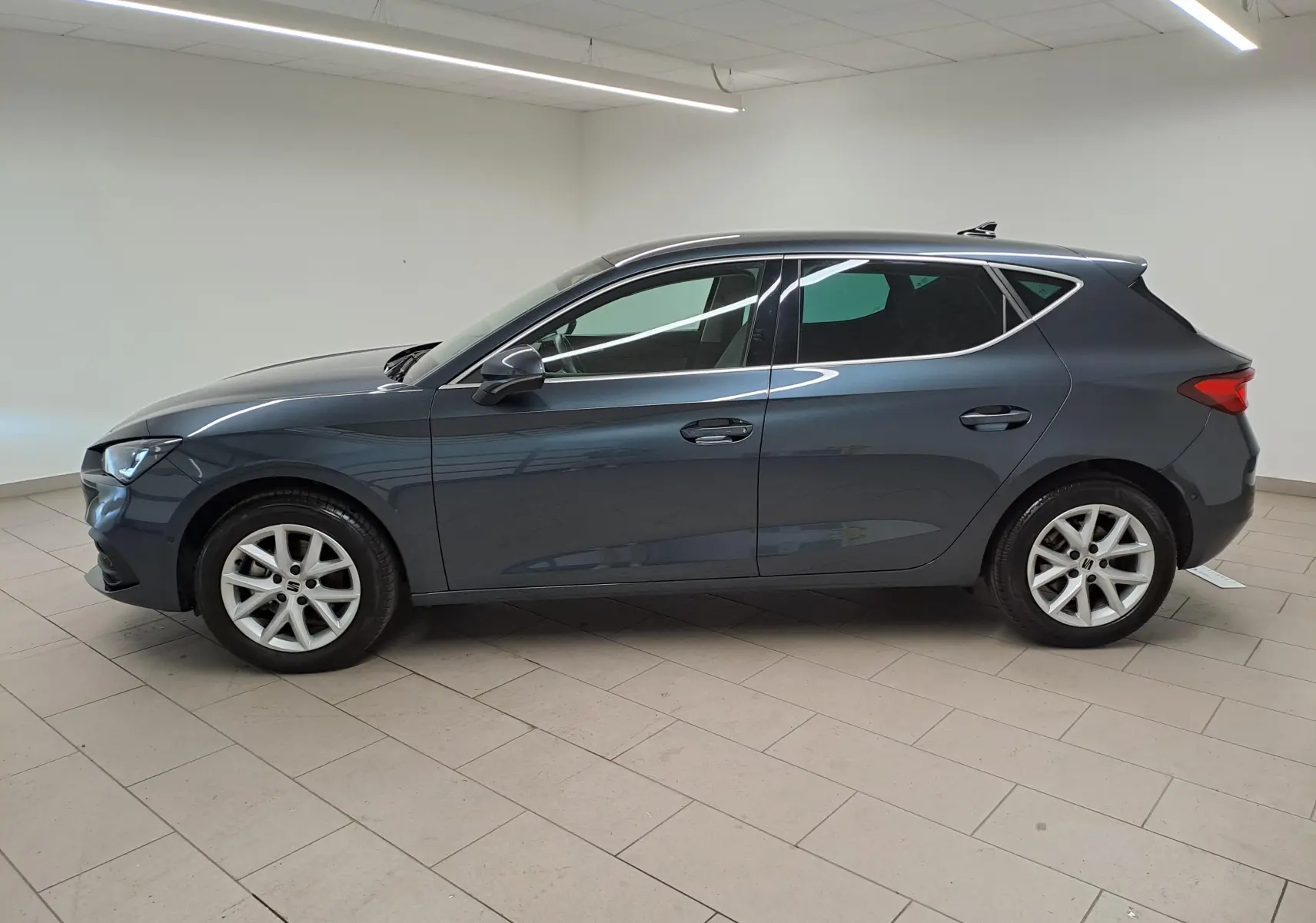Vue de profil côté gauche d'une SEAT LEON 1.5 eTSI Hybrid gris magnétique avec vitres arrière surteintées en intérieur neutre.