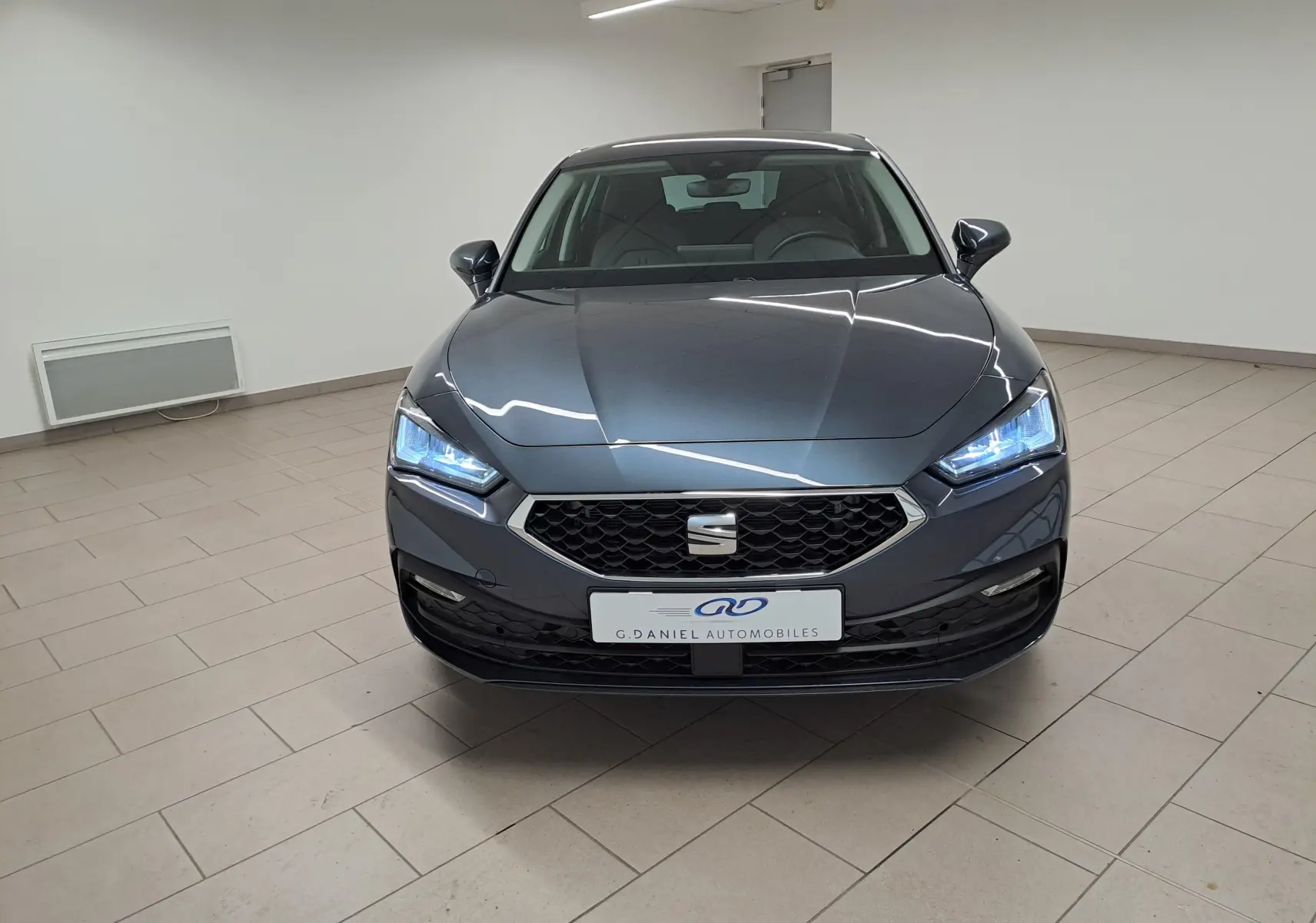 Vue de face d'une SEAT LEON 1.5 eTSI Hybrid gris magnétique avec phares allumés dans un intérieur neutre.