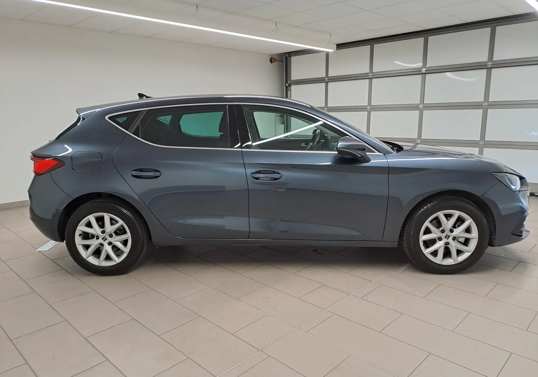 Vue de profil côté gauche d'une SEAT LEON 1.5 eTSI Hybrid gris magnétique avec vitres arrière surteintées en intérieur showroom.