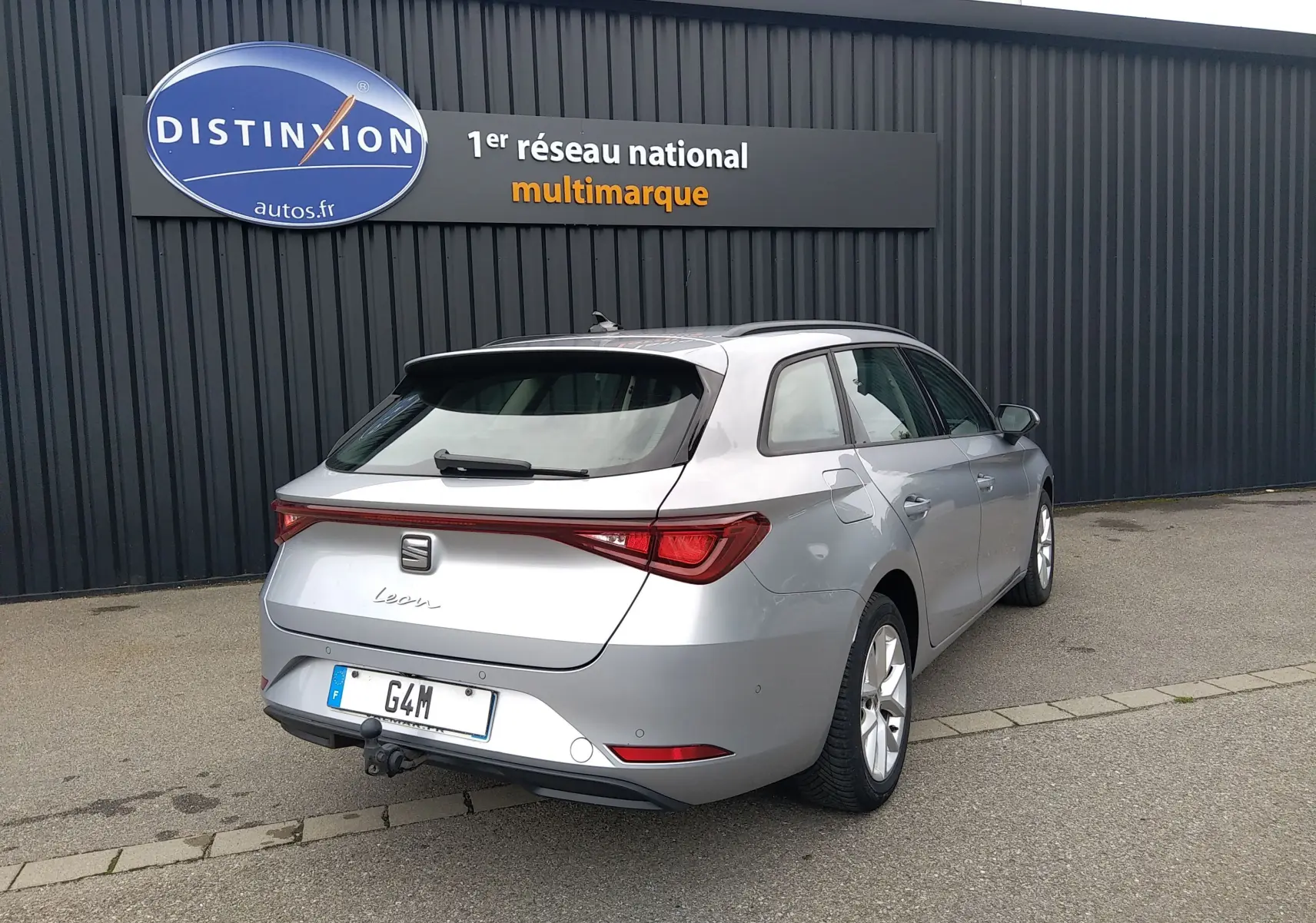 SEAT Leon ST gris vue 3/4 arrière droit avec attelage, stationnée devant un mur noir et un panneau Distinxion.