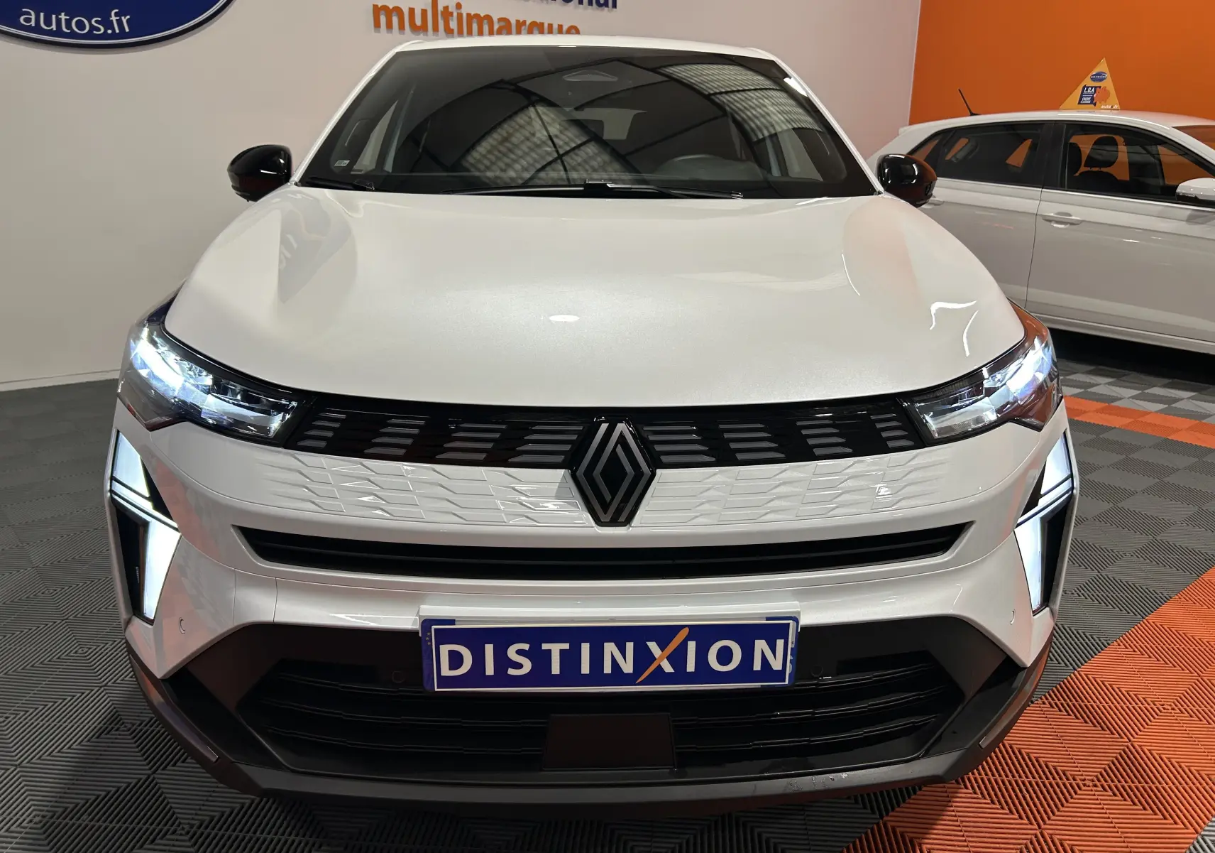 Vue frontale d'une Renault Symbioz blanc nacré avec phares LED allumés et calandre moderne en intérieur showroom.