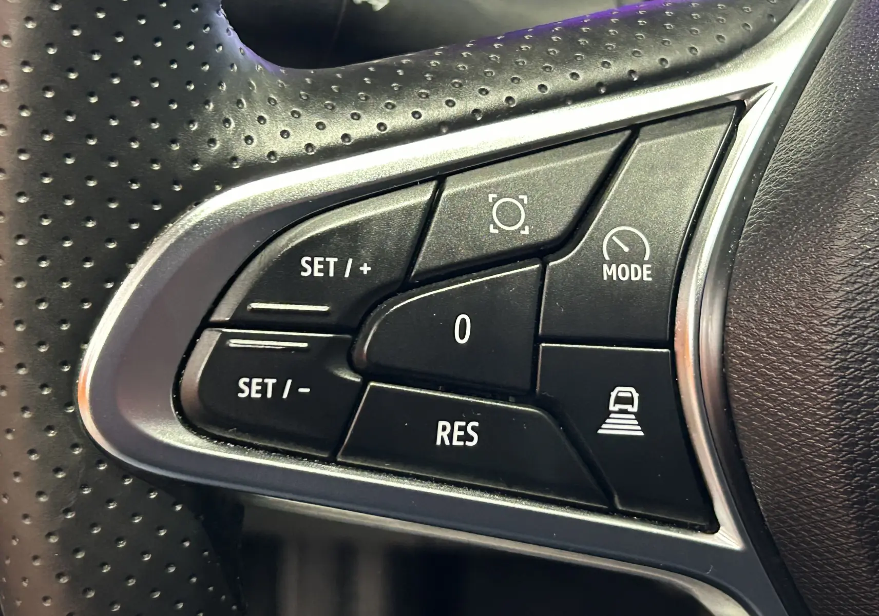 Gros plan sur les commandes noires du volant perforé de la Renault Symbioz blanc nacré, boutons de régulateur et mode conduite.