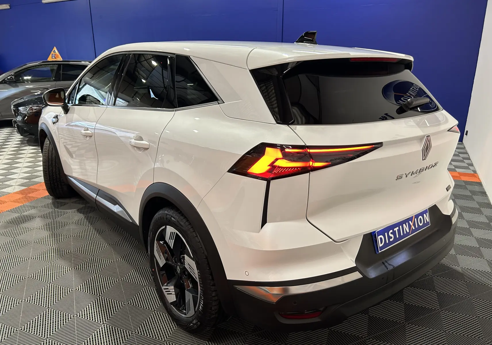 Vue 3/4 arrière droite d'un Renault Symbioz blanc nacré avec feux arrière LED allumés en intérieur showroom.