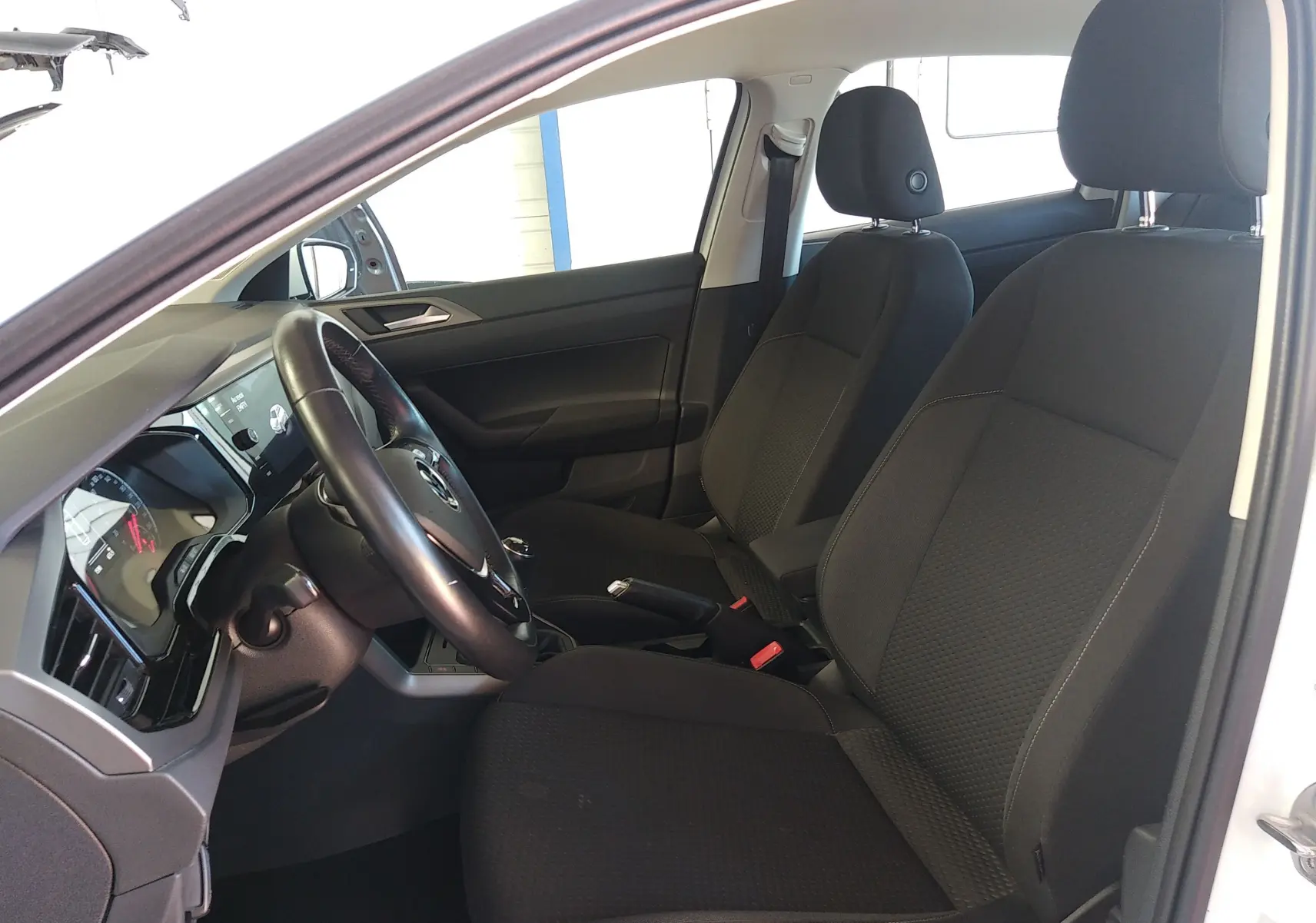 Vue intérieure côté conducteur de la Volkswagen Polo blanche 2021, montrant les sièges noirs et le tableau de bord moderne.