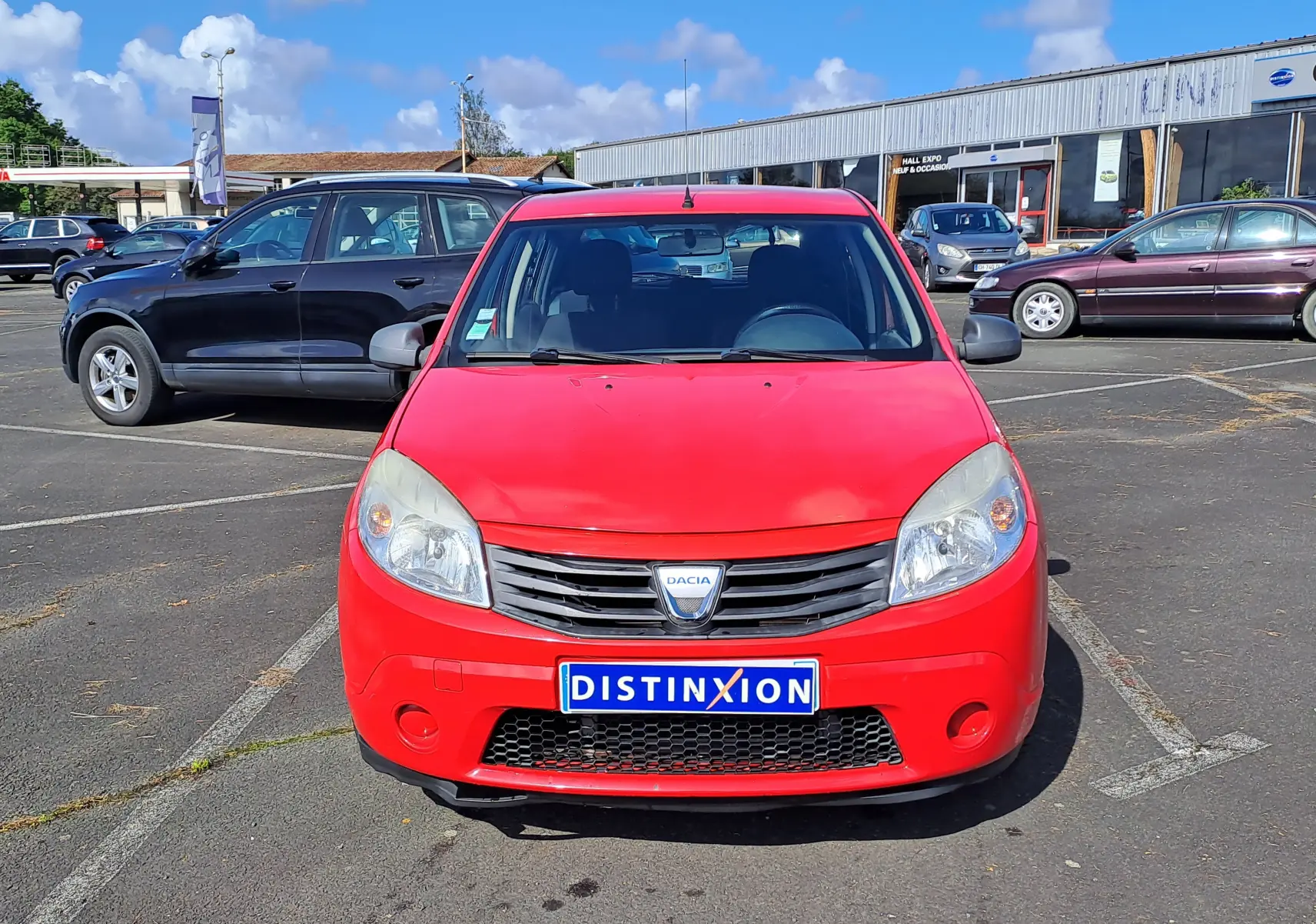 Vue de face d'une Dacia Sandero rouge de 2010 avec plaque Distinxion, stationnée sur un parking extérieur.