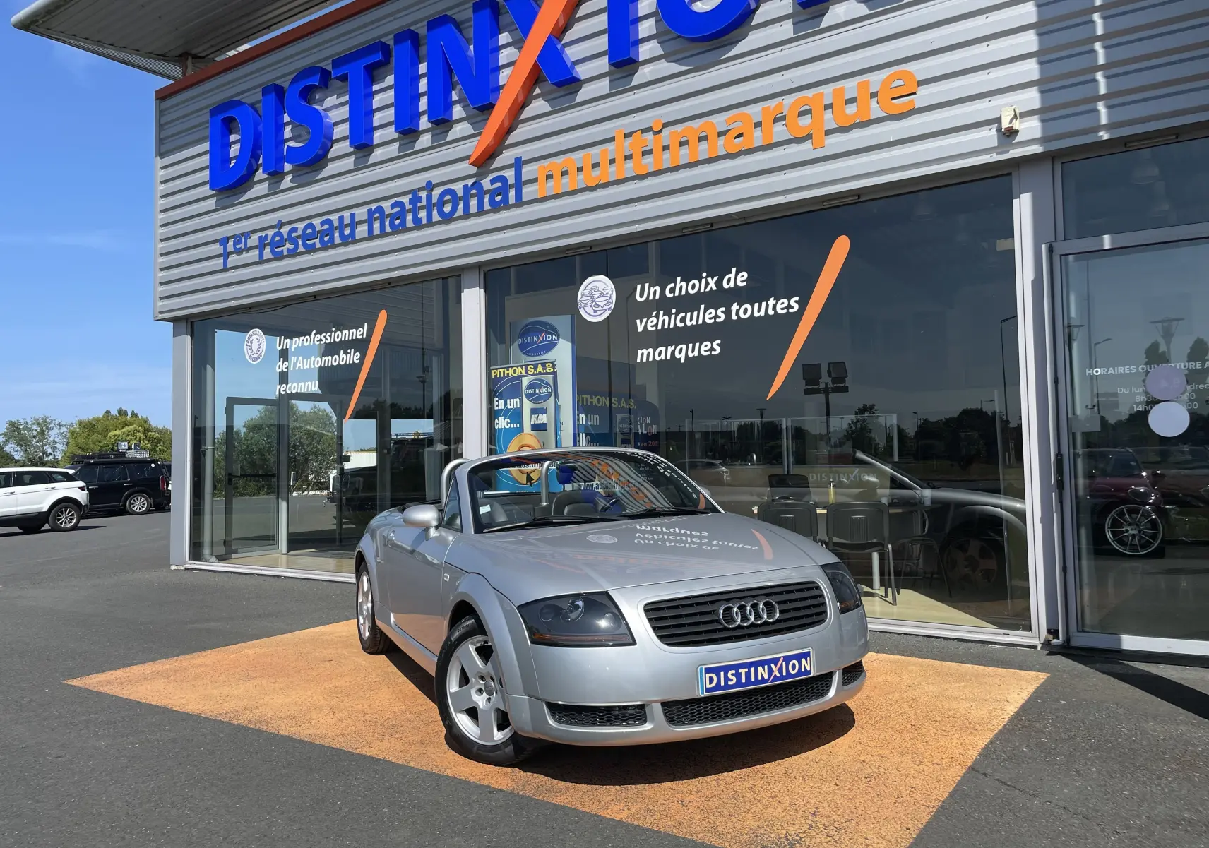 Audi TT Roadster 1.8T 180 gris argent vu en 3/4 avant, capote ouverte, stationné devant un showroom Distinxion.