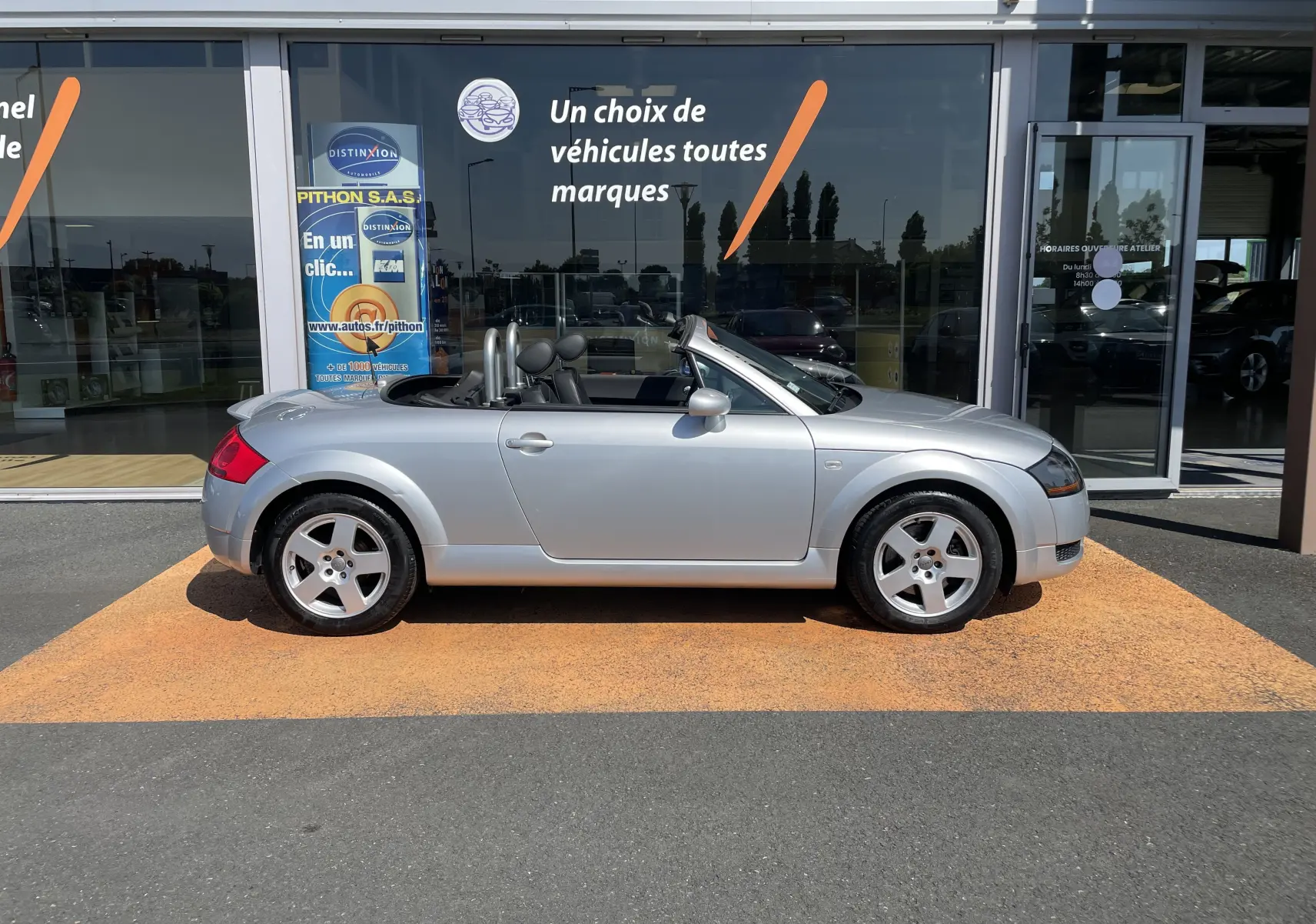 Profil côté gauche d'un Audi TT Roadster gris argent 2002 décapoté, avec jantes 5 branches et intérieur noir visible.