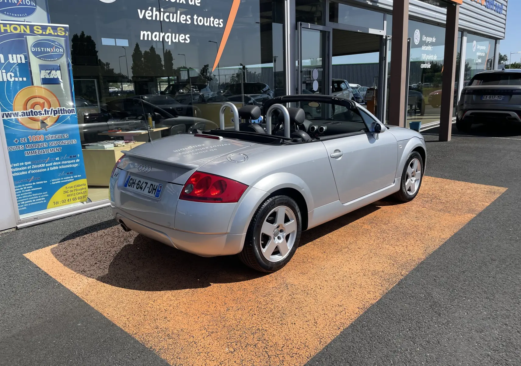 Audi TT Roadster 1.8T 180 gris argent vu de 3/4 arrière droit, capote ouverte, stationné devant un showroom.