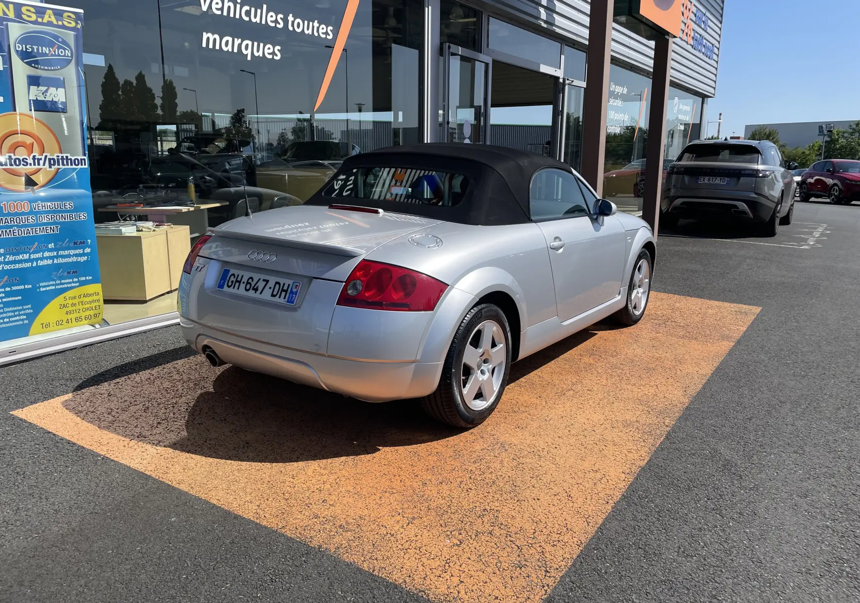 Audi TT Roadster gris argent 2002 vue 3/4 arrière droit avec capote noire, stationné devant un showroom.