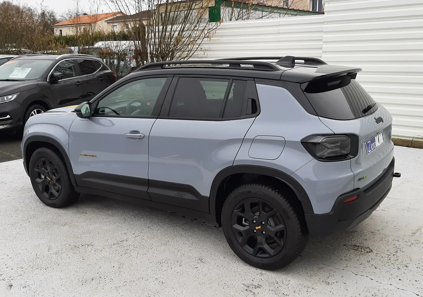 Jeep Avenger 2025 gris Storm avec toit noir, vue 3/4 arrière côté gauche, jantes noires et barres de toit.