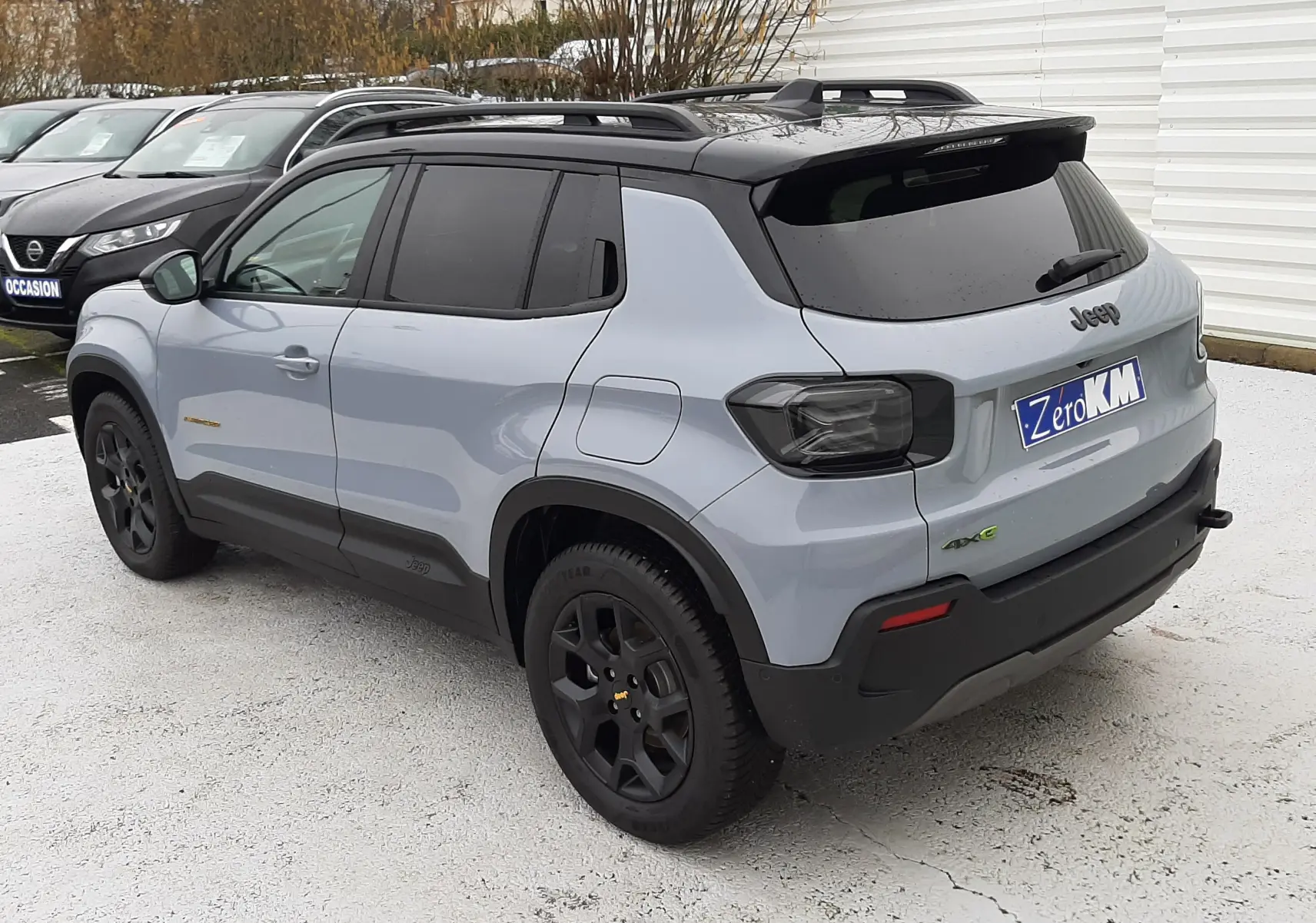Jeep Avenger 2025 gris Storm avec toit noir, vue 3/4 arrière côté gauche, jantes noires et feux arrière fumés.
