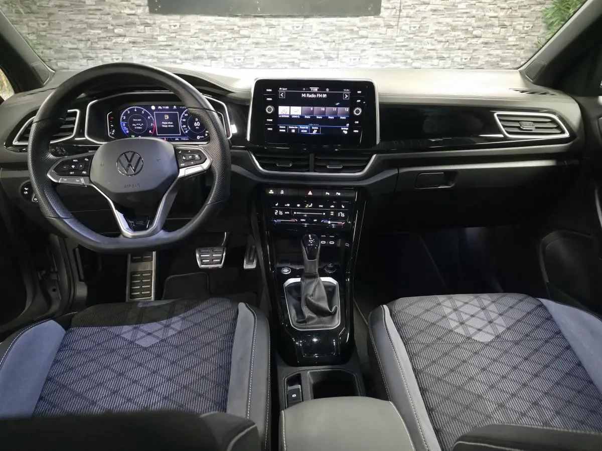 Vue intérieure avant du Volkswagen T-Roc 1.5 TSI R-Line 2025, avec tableau de bord digital et sièges tissu gris à motifs.