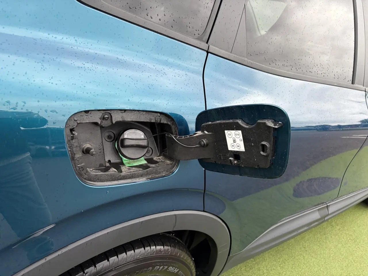 Gros plan sur la trappe à carburant ouverte côté gauche d’un Renault Arkana bleu Zanzibar métal sous la pluie.