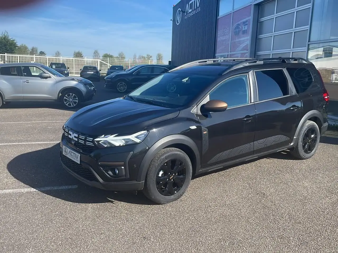 Dacia Jogger noir nacré en 3/4 avant droit avec jantes noires et barres de toit visibles sur parking.