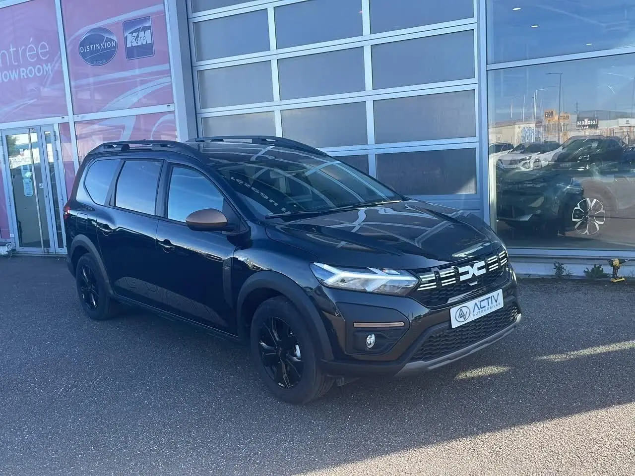 Dacia Jogger noir nacré en 3/4 avant droit avec barres de toit et jantes noires devant un showroom.