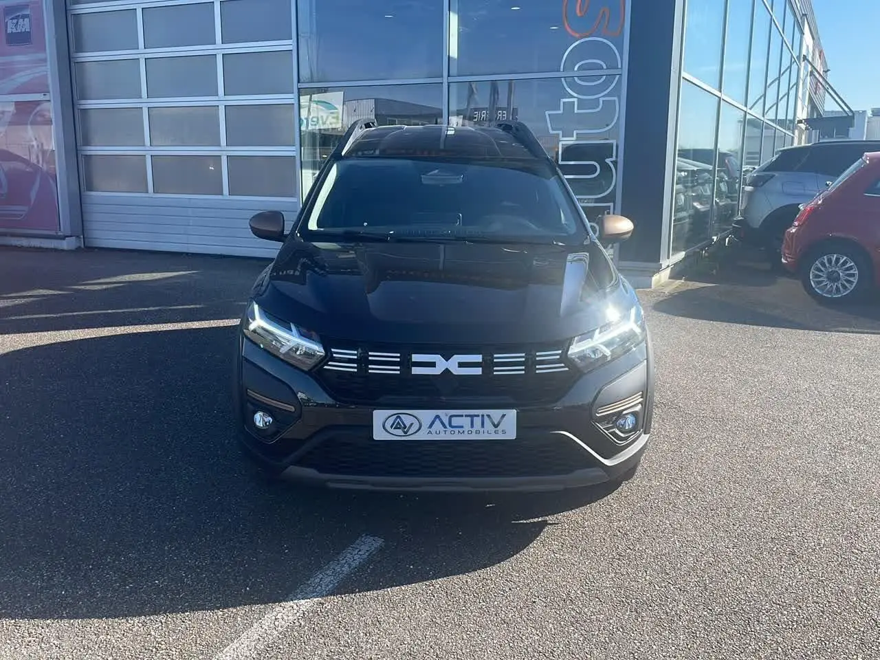 Vue de face du Dacia Jogger noir nacré avec calandre distinctive et phares LED allumés, stationné devant un garage.