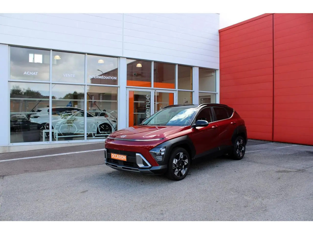 Hyundai Kona 2024 rouge Ultimate Metal avec toit noir, vu en 3/4 avant droit devant un bâtiment moderne.