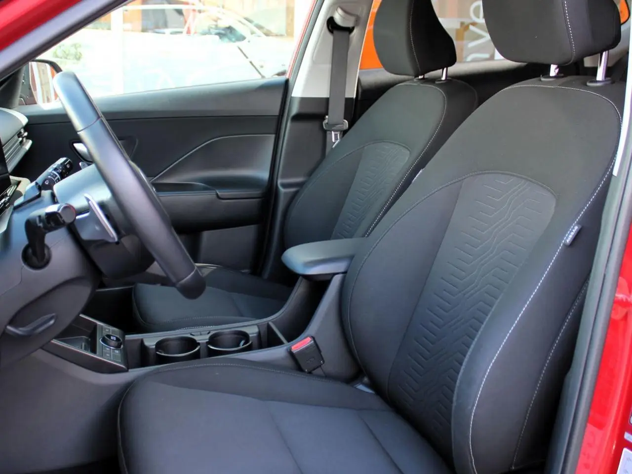Vue intérieure côté conducteur du Hyundai Kona 2024 rouge, sièges avant noirs avec accoudoir central.