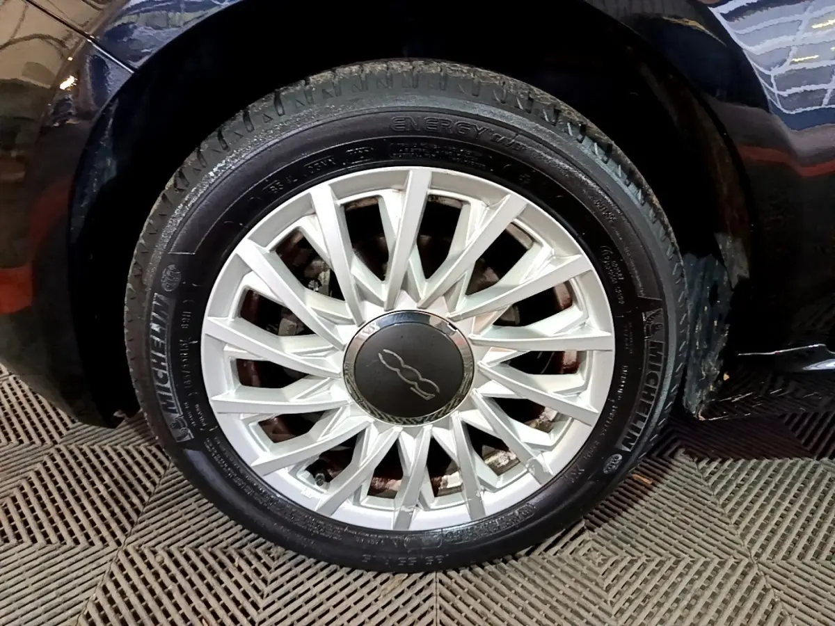 Gros plan sur la roue avant droite d'une FIAT 500 noire, mettant en valeur la jante argentée au design étoilé.