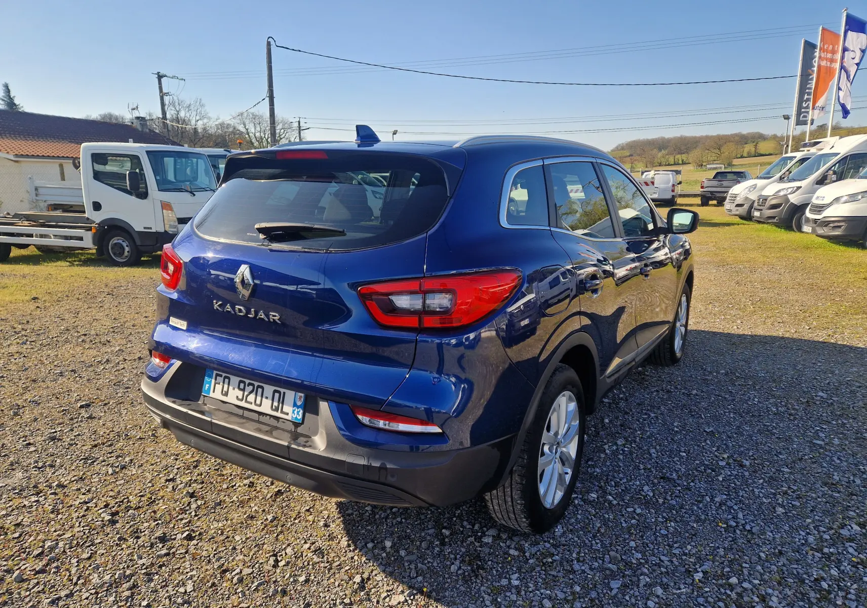 Vue 3/4 arrière droite d’un Renault Kadjar bleu cosmos 2020 garé sur un sol gravillonné en extérieur.