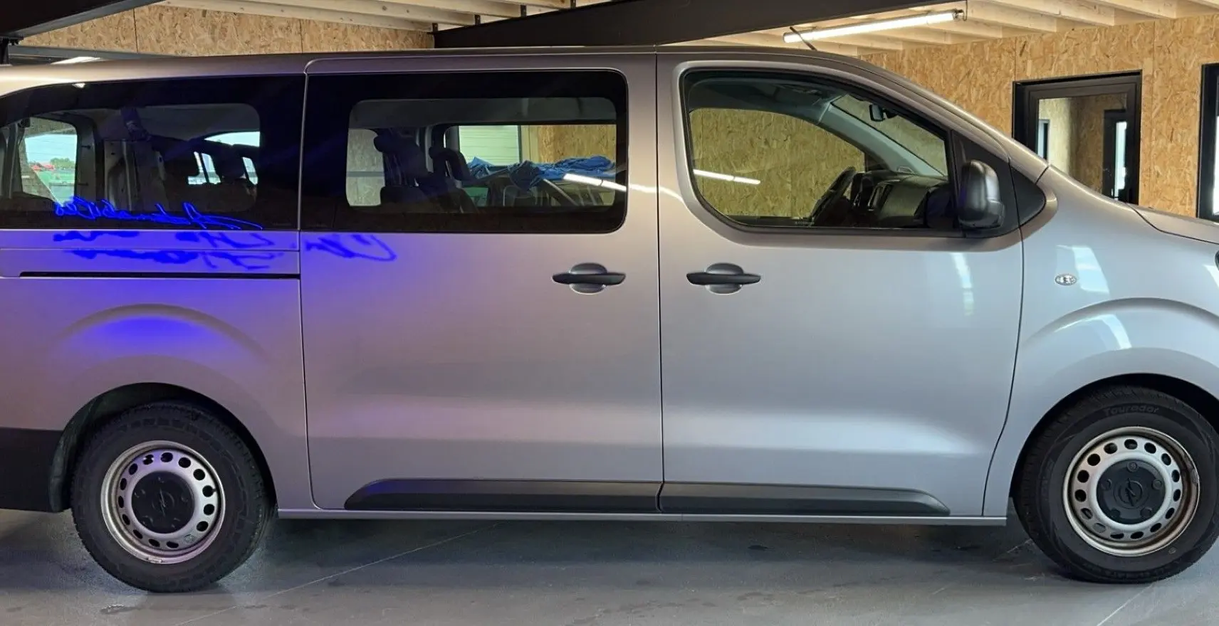 Profil côté gauche d'un Opel Vivaro Combi gris 2022 avec vitres teintées et jantes acier en intérieur d'atelier.