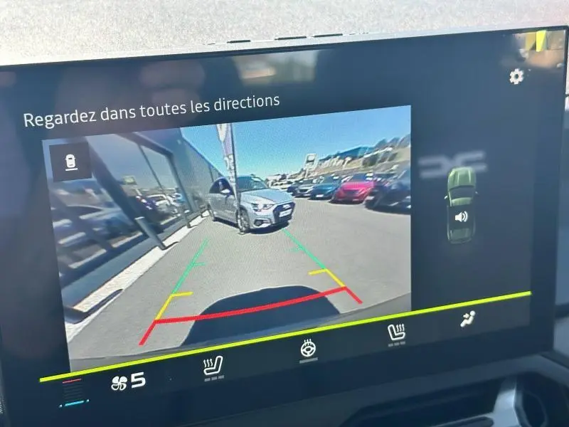 Image de la caméra de recul montrant un Dacia Duster blanc en 3/4 avant dans un parking extérieur ensoleillé.