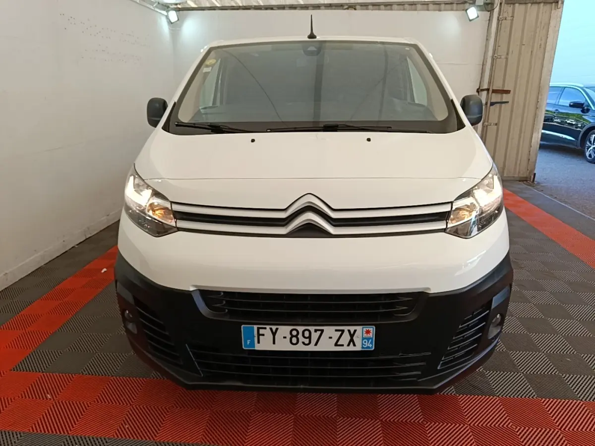 Vue frontale d'un Citroën Jumpy XS blanc avec pare-chocs noir et phares allumés en intérieur.