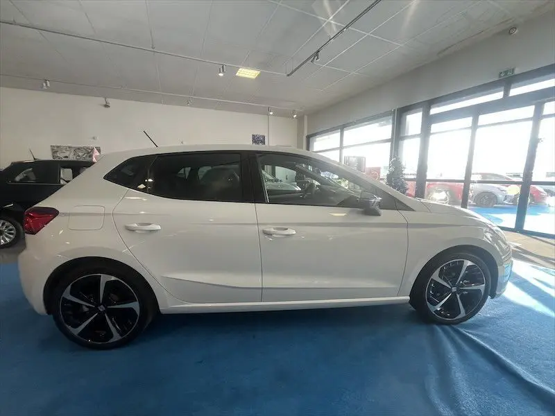 Profil côté gauche d'une SEAT Ibiza 2024 blanc Nevada métal avec jantes noires et toit noir en showroom lumineux.