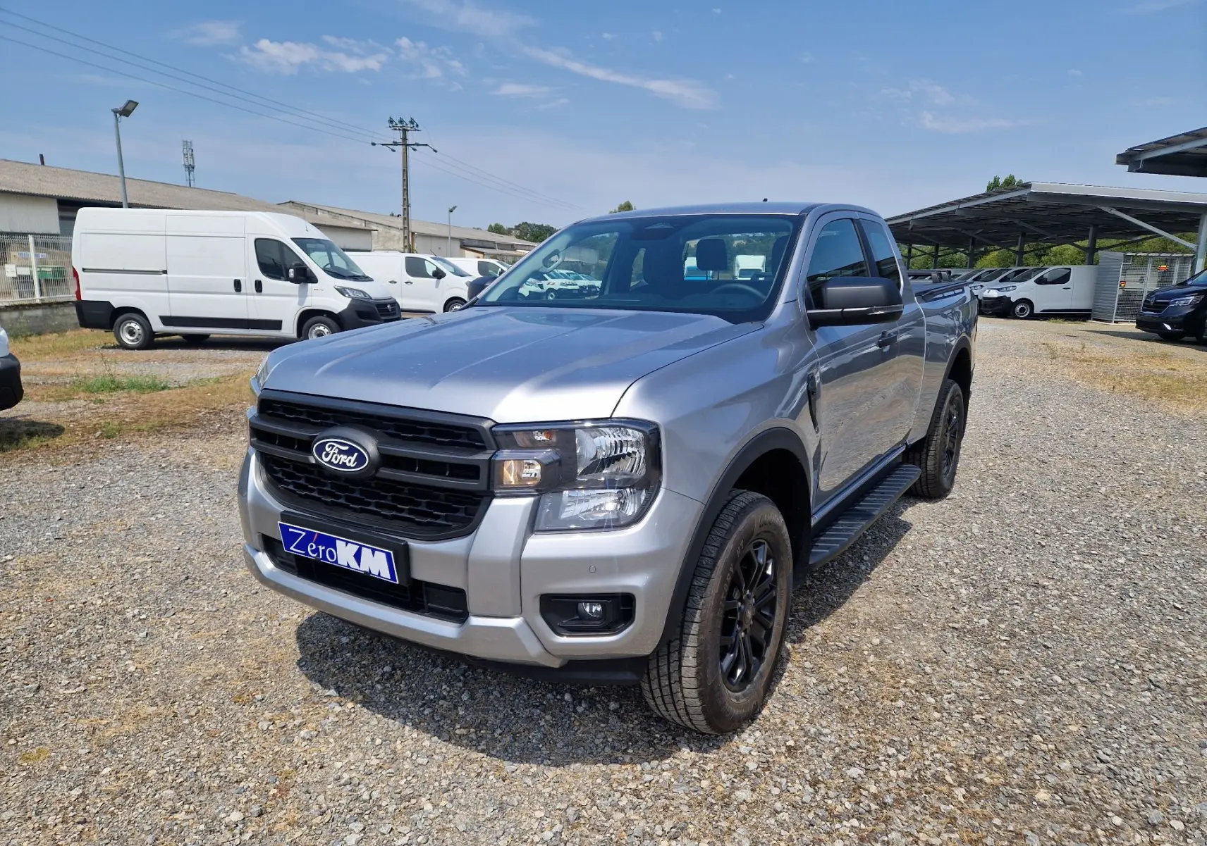 Ford Ranger Super Cabine gris Iconic en 3/4 avant droit, avec marchepied noir et jantes foncées sur terrain gravillonné.