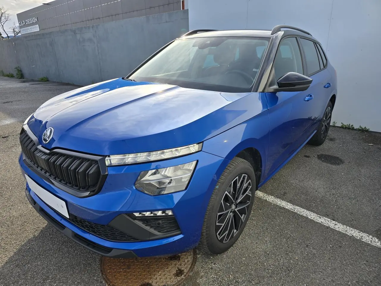 Vue 3/4 avant d’une Skoda Kamiq bleu racing avec toit noir et jantes alliage 17 pouces, stationnée sur un parking.