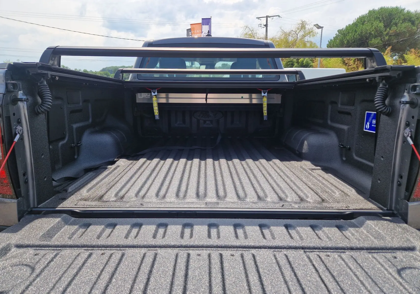 Vue arrière du plateau ouvert d'un Ford Ranger Super Cabine gris carbone, avec protection renforcée et barres de chargement visibles.