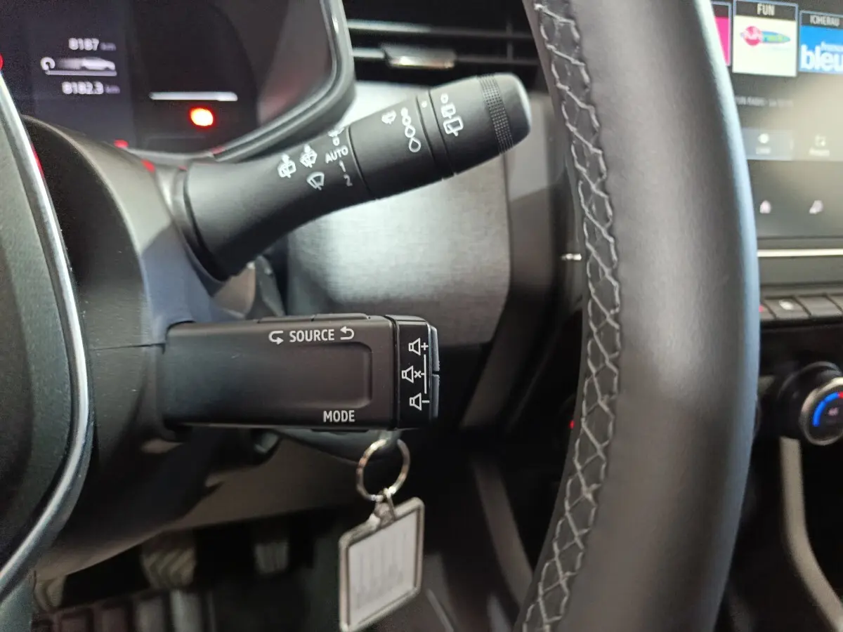 Gros plan sur le volant cuir synthétique et les commandes au volant d’une Renault Clio gris clair, tableau de bord partiellement visible.