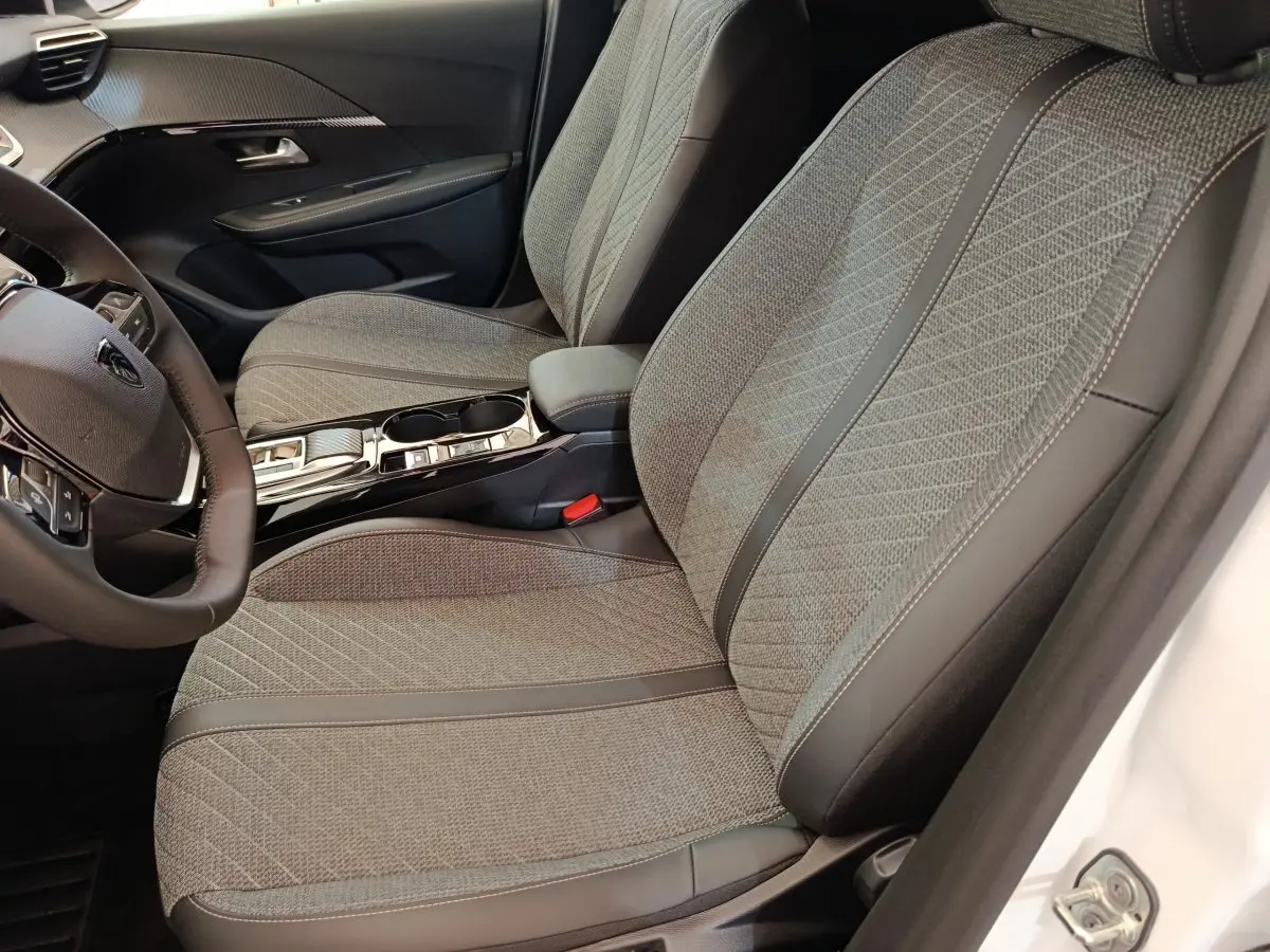 Vue intérieure côté conducteur de la Peugeot 208 électrique blanche 2024, sièges tissu gris avec surpiqûres et volant cuir.