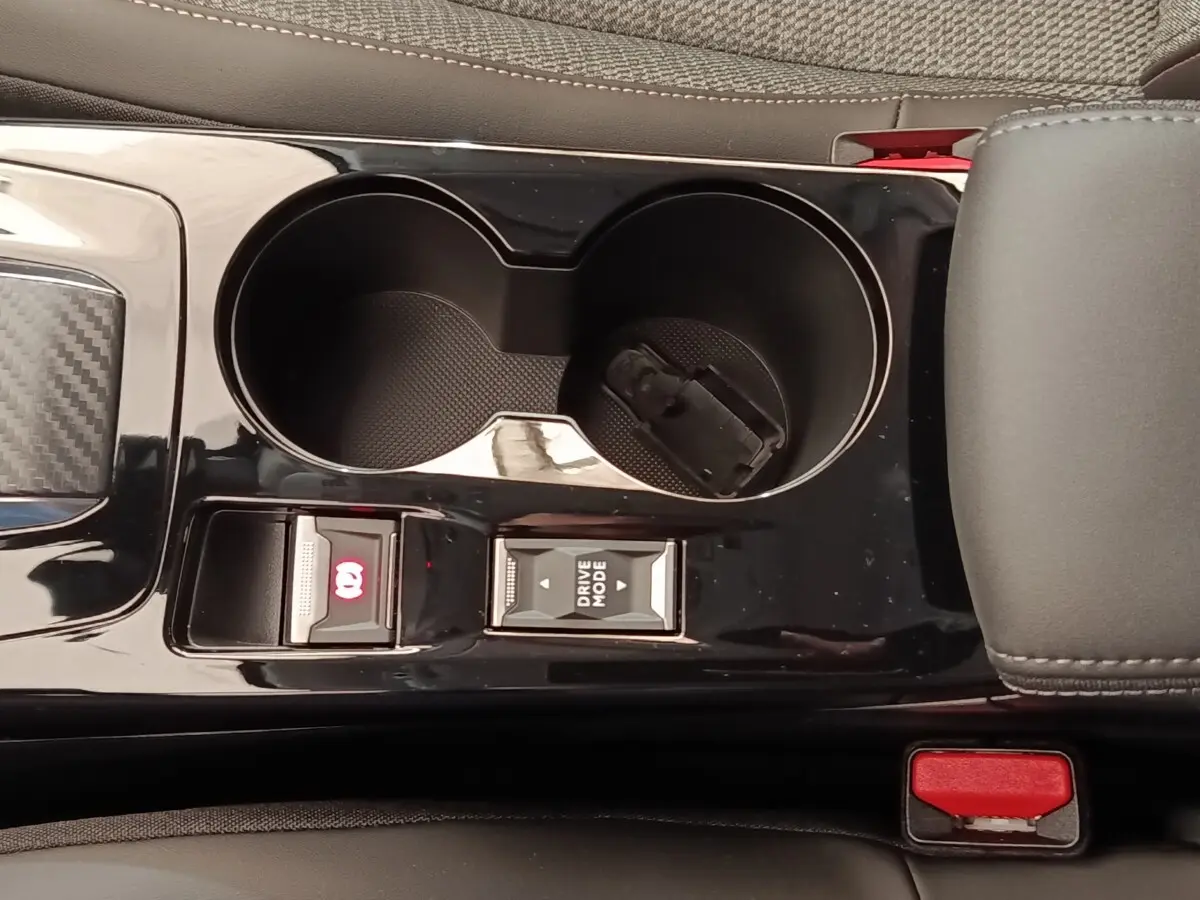 Gros plan sur la console centrale noire de la Peugeot 208 électrique 2024, avec porte-gobelets et commandes frein à main et mode de conduite.