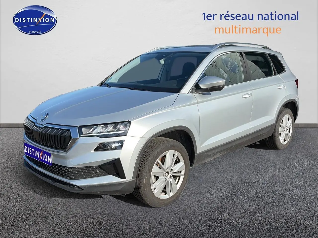 Skoda Karoq gris 3/4 avant droit, avec calandre noire et jantes alliage visibles sur sol gris.